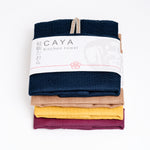 A stack of four, different coloured, folded muslin Caya kitchen towels, made in Japan using traditional mosquito-net weaving techniques. Each tea towel has a small braided loop that shows under the white, paper packaging label wrapped around each one. Colours from top to bottom: navy blue, brown, yellow and grape burgundy. Available at NiMi Projects UK.