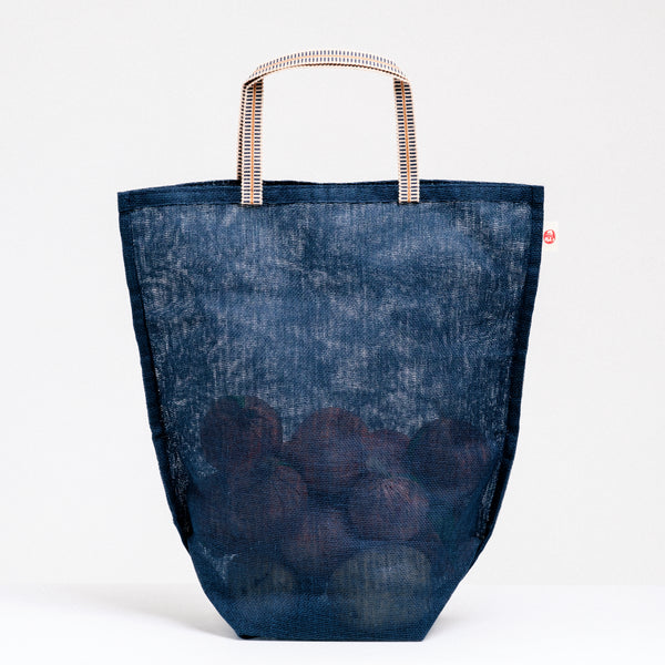 A large navy blue muslin Vegetable tote bag filled with onions, made in Japan by Ban Inoue using traditional mosquito net weaving techniques. The bag has a folded and stitched base for support and short top handles. On show at NiMi Projects UK.