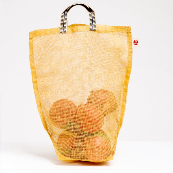 A medium yellow muslin Vegetable tote bag filled with several onions, made in Japan by Ban Inoue using traditional mosquito net weaving techniques. The bag has a folded and stitched base for support and short top handles. On show at NiMi Projects UK.