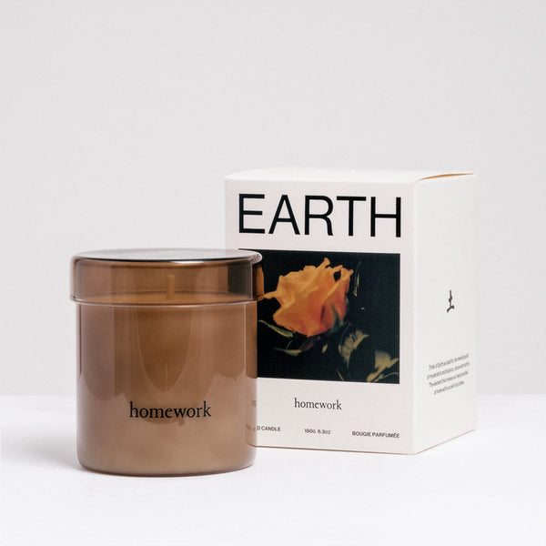 A Homework Earth-scented white candle inside a brown tinted jar, standing in front of the Homework branded box. Available at NiMi Projects UK.