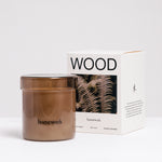 A Homework Wood-scented white candle inside a brown tinted jar, stading in front of the Homework branded box. Available at NiMi Projects UK.
