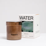 A Homework Water-scented white candle inside a brown tinted jar, standing in front of the Homework branded box. Available at NiMi Projects UK.