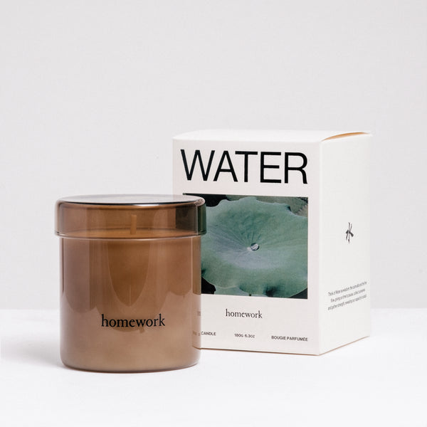 A Homework Water-scented white candle inside a brown tinted jar, standing in front of the Homework branded box. Available at NiMi Projects UK.