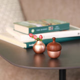 A small singing bowl chime made of pink brass, featuring a wooden like shaped like an acorn cap. The bell is placed on a dark brown side table with a small pink brass bud vase.