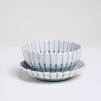 A white, Japanese porcelain, Mino-yaki, pinstriped, bean-bowl dish sitting on top of a matching small plate. Both items feature a gently fluted edge and radial lines in blue underglaze. On show at NiMi Projects UK.