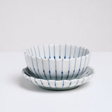 A white, Japanese porcelain, Mino-yaki, pinstriped, bean-bowl dish sitting on top of a matching small plate. Both items feature a gently fluted edge and radial lines in blue underglaze. On show at NiMi Projects UK.