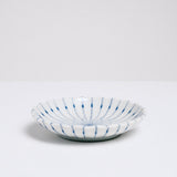 A side view of a white, Japanese porcelain, Mino-yaki pinstriped side dish, with a gently fluted edge and radial hairline thin lines in blue underglaze. On show at NiMi Projects UK.