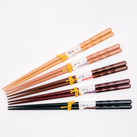 Five pairs of wooden chopsticks, laid fanned out, each with a carved indented pattern at the top and made from a different wood or with a different finish, and all available  from NiMi  Projects UK. From  top to bottom: Tsuge boxwood, Buna beech, Tsuge ironwood, Shitan rosewood finish and Kuroban blackwood finish.