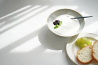 A portion of yoghurt served in a Kihon no Utsuwa Japanese porcelain bowl in white, with a small side plate by the same ceramic brand holding bread and grapes.