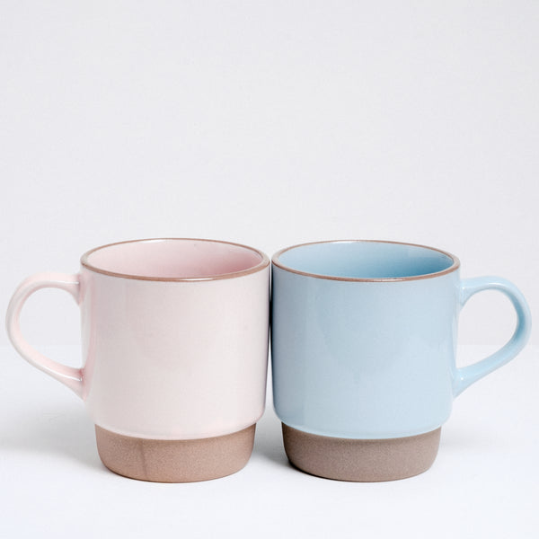 A pair of NiMi Projects’ Japanese ceramic Mino-yaki stacking mugs — one pink and one blue — placed side by side. Each mug features a pastel satin glaze and has an unglazed base and lip.