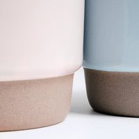 A detail image of a pair of NiMi Projects’ Japanese ceramic Mino-yaki stacking mugs — one pink and one blue — placed side by side and showcasing each mug’s pastel satin glaze and unglazed base.