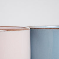 A detail image of the tops of two NiMi Projects’ Japanese ceramic Mino-yaki stacking mugs — one pink and one blue — placed side by side and showcasing each mug’s pastel satin glaze and unglazed lip.