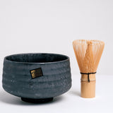 A black, ceramic Japanese matcha tea chawan bowl with a bamboo chasen tea whisk to its right. The bowl is lightly textured with ridges and features the maker's gold brand sticker, and its glaze accented with a subtle shimmer. Available at NiMi Projects UK.