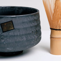 A NiMi Projects' closeup image of a black, ceramic Japanese matcha tea chawan bowl with a bamboo chasen tea whisk to its right. The bowl features the maker's sticker and is lightly textured with ridges and its glaze accented with a subtle shimmer.