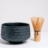 A black, ceramic Japanese matcha tea chawan bowl with a bamboo chasen tea whisk to its right. The bowl is lightly textured with ridges and its glaze accented with a subtle shimmer. Available at NiMi Projects UK.