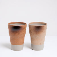 A pair of Mino-yaki., Shigaraki style, Japanese ceramic tumblers, both featuring a raw ceramic base and glazed above with a speckled and naturally singed pattern. On the lefft is a soft orange tumbler; on the right a creamy yellow tumbler. Available at NiMi Projects UK.