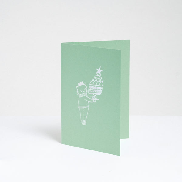 A standing pistachio green greeting card, designed by Takako Copeland, featuring a white line drawing illustration of a cat in a party crown and sweater holding a large trifle on a platter. Available at NiMi Projects UK. 