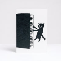 A letterpress printed greeting card by Takako Copeland, featuring a top view of a black grand piano and two black illustrated cats playing at its sides.