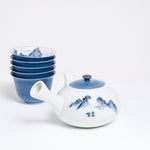 A vintage white porcelain Japanese tea set, including a tea yokode (side-handled) teapot and five stacked teacups. The teapot features a blue lid and a hand-painted blue design of a mountainscape, while the stacked cups have blue exteriors and mountains painted inside. Available at NiMi Projects UK.