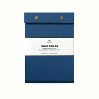 A lake blue pressed-cotton covered Postalco Snap Pad, showing the two roundmetal snap clasps at its top and a white label wrapped around its center featuring the Postalco bird logo.