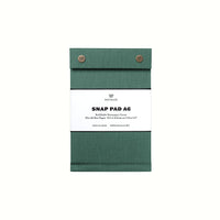 A dark green pressed-cotton covered A6 Postalco Snap Pad, showing the two roundmetal snap clasps at its top and a white label wrapped around its center featuring the Postalco bird logo.