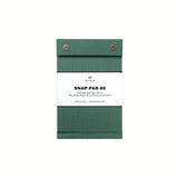 A dark green pressed-cotton covered A6 Postalco Snap Pad, showing the two roundmetal snap clasps at its top and a white label wrapped around its center featuring the Postalco bird logo.