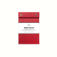 A signal-red fabric-covered A6 Postalco Snap Pad, showing the two roundmetal snap clasps at its top and a white label wrapped around its center featuring the Postalco bird logo.