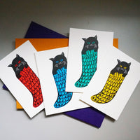 A group of four Takako Copeland illustrated and letterpress printed Christmas cards, featuring black cats in different coloured knitted stockings. From left to right: a black cat in a red stocking, blue stocking, green stocking and yellow stocking.